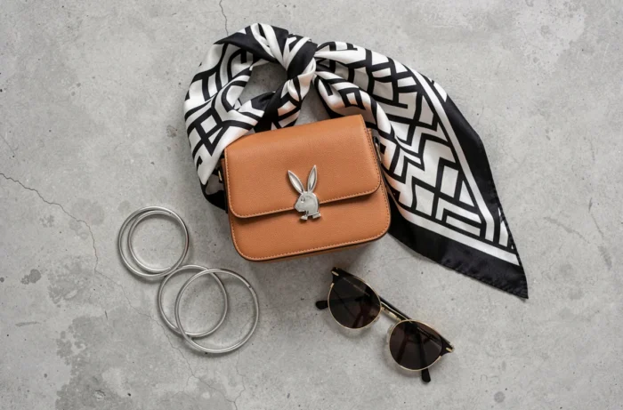 Flat-lay éditorial d'accessoires tendance inspirés par Playboy incluant sac en cuir, foulard en soie et bijoux dorés